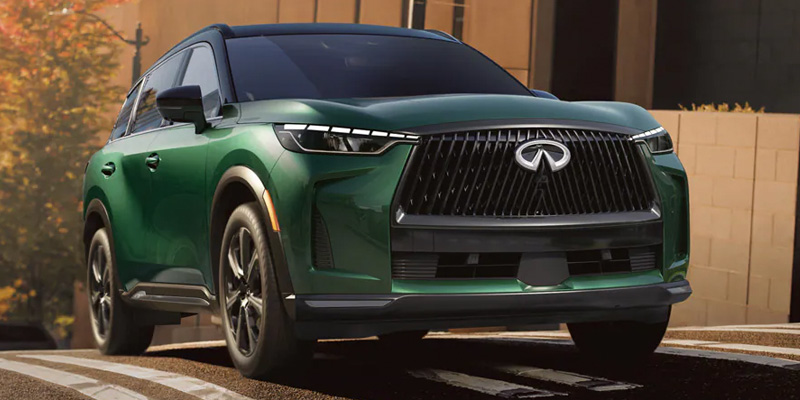 Green Infiniti QX60 SUV driving on an urban road with modern buildings in the background.